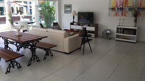 A spacious living area with a sofa, wooden table, and chairs. A television is mounted on the wall, and there is a fan on the floor. - Beira Mar House in Angra dos Reis - Close to commerce in secure condominium (Angra dos Reis)