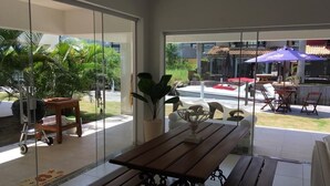 A spacious patio area with a wooden table and benches, surrounded by glass doors leading to a garden with palm trees and a view of a restaurant with outdoor seating. - Beira Mar House in Angra dos Reis - Close to commerce in secure condominium (Angra dos Reis)