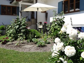 A garden with various plants and flowers, a patio area with a table and chairs, and a white umbrella. - Apartment for 2 persons, 50sqm, ground floor with terrace (Bad Feilnbach)