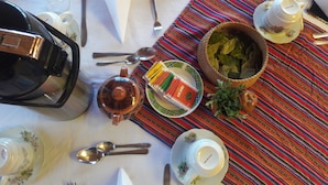 Una mesa servida con tetera, tazas, cucharas, un tazón de hierbas y un mantel individual colorido.