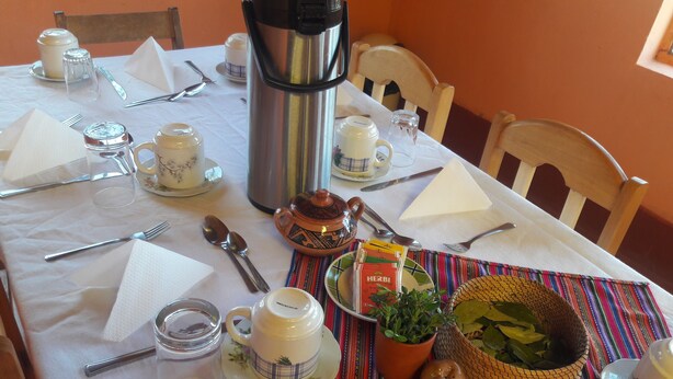 Una mesa de comedor dispuesta con varios utensilios, un termo, una tetera, un plato con una caja de té 'Herbi', una planta en maceta y una cesta tejida.