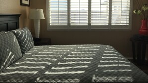 Une chambre à coucher avec un lit, une fenêtre avec des stores, une lampe de chevet et un tableau encadré au mur.