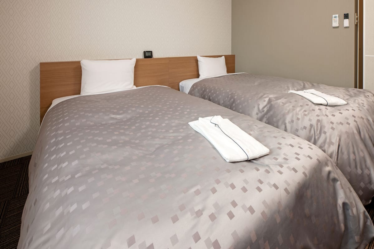 Habitación de hotel con dos camas, cada una con cabecero de madera y ropa de cama estampada. Se observa un panel de control en la pared y una puerta a la derecha.