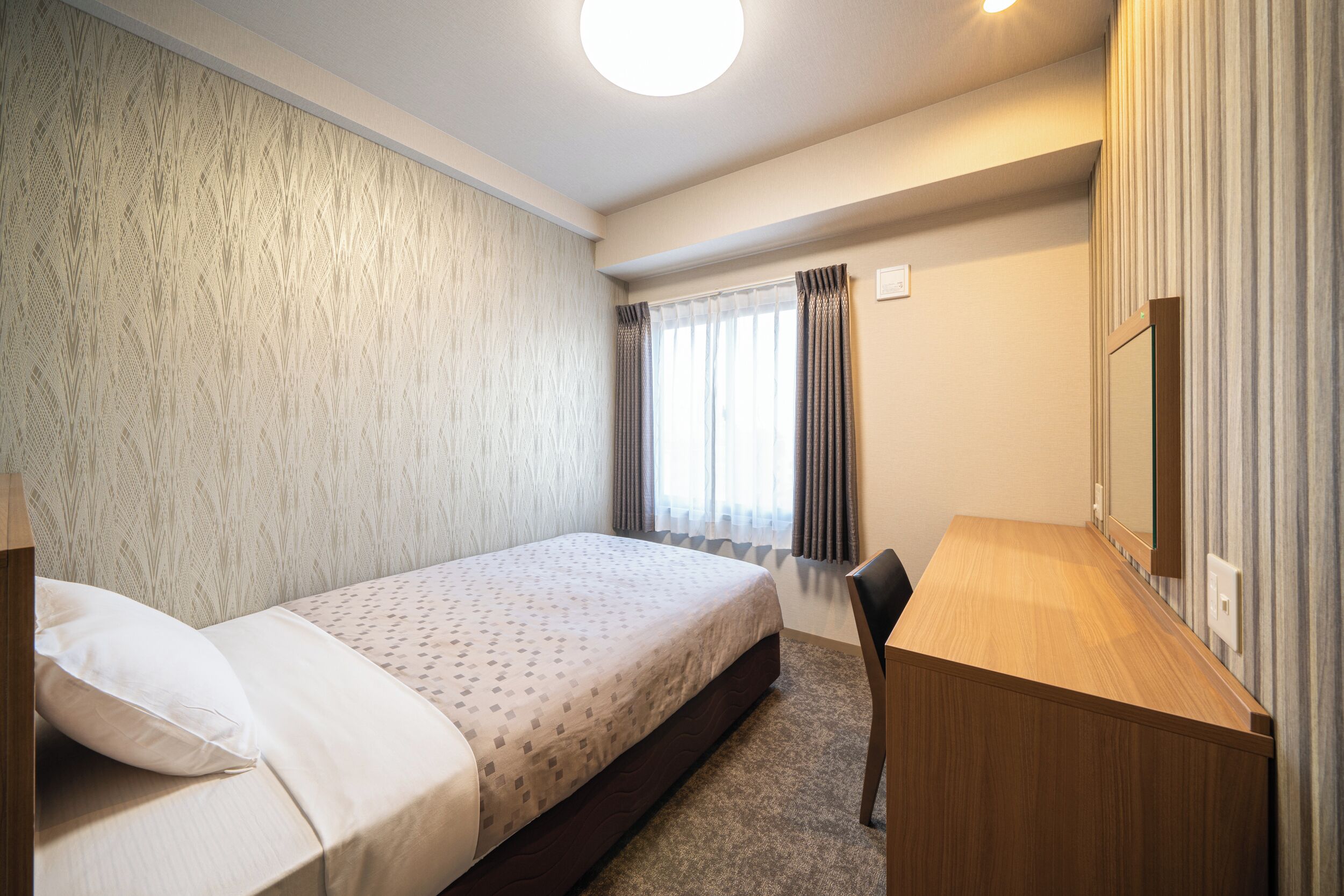 Hotellihuone, jossa on sänky, työpöytä, tuoli, ikkuna verhoilla ja seinälle kiinnitetty peili.