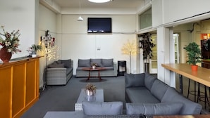 Een moderne woonkamer met een grijze bank, een kleine ronde tafel en een aan de muur bevestigde televisie. In de achtergrond staan planten in potten en een houten bar.