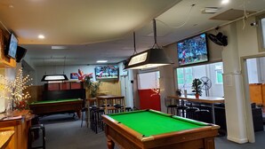 Een poolzaal met meerdere pooltafels, een bar en een televisie aan de muur.