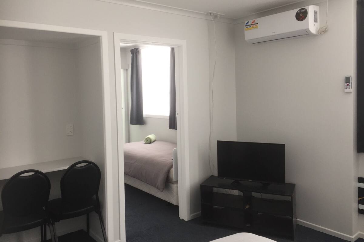 A small, sparsely furnished room with a bed, a desk with chairs, a television, and an air conditioning unit.