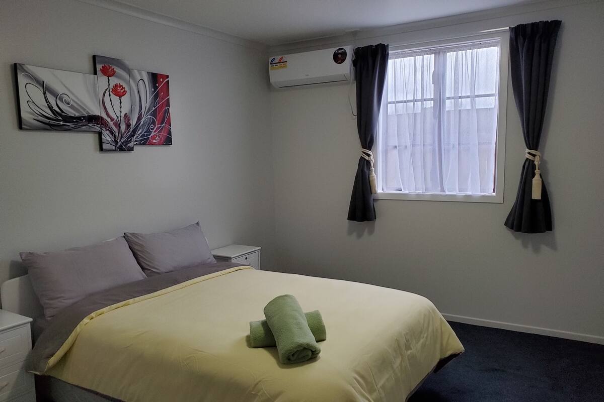 A hotel room with a bed, a nightstand, an air conditioning unit, and a window with curtains.
