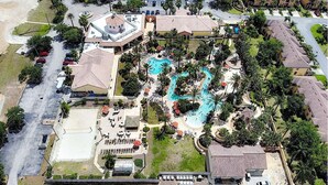 An aerial view of a resort with multiple swimming pools, a large parking area, and surrounding residential buildings.