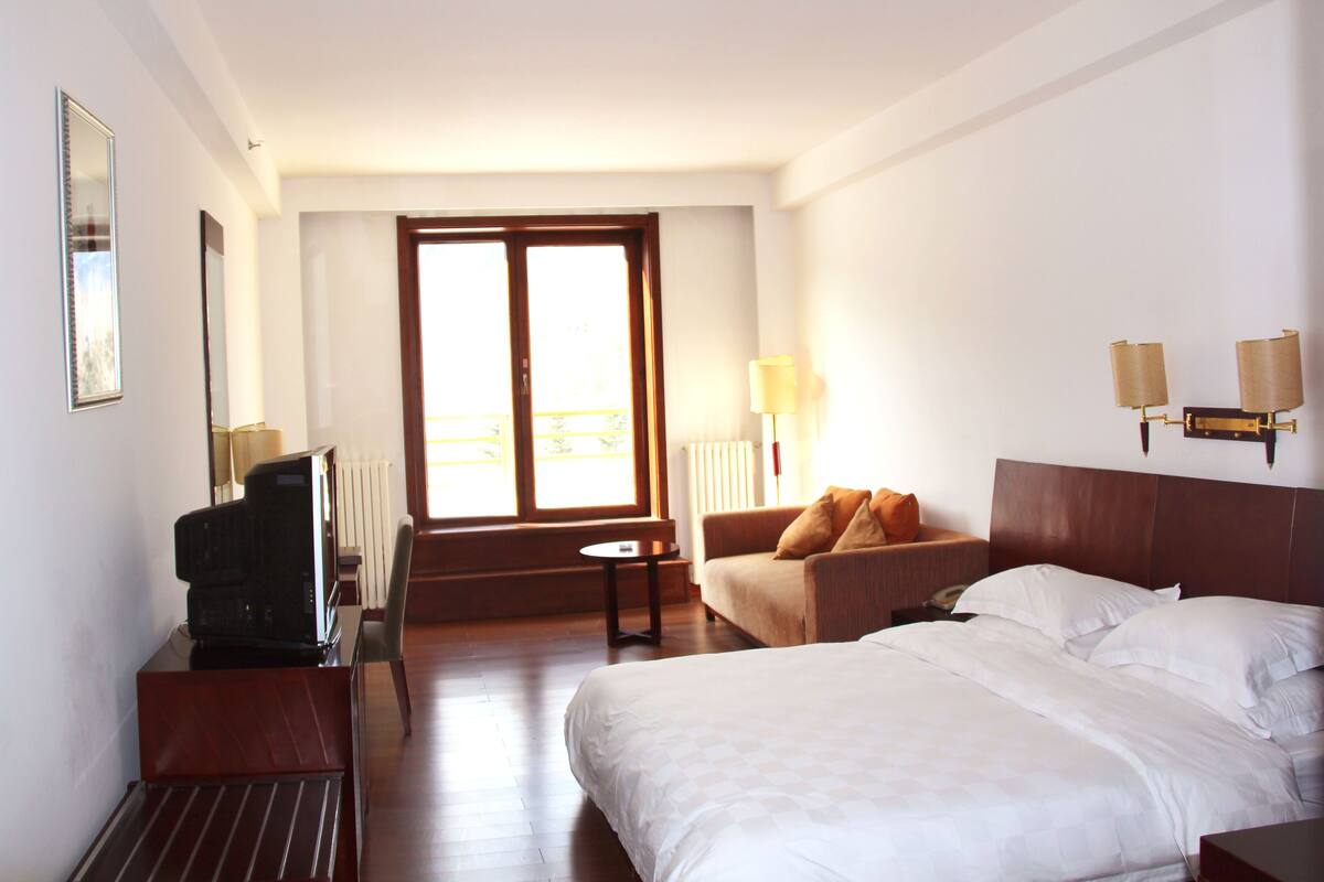 A hotel room with a bed, a sofa, a TV, and a window with blinds.