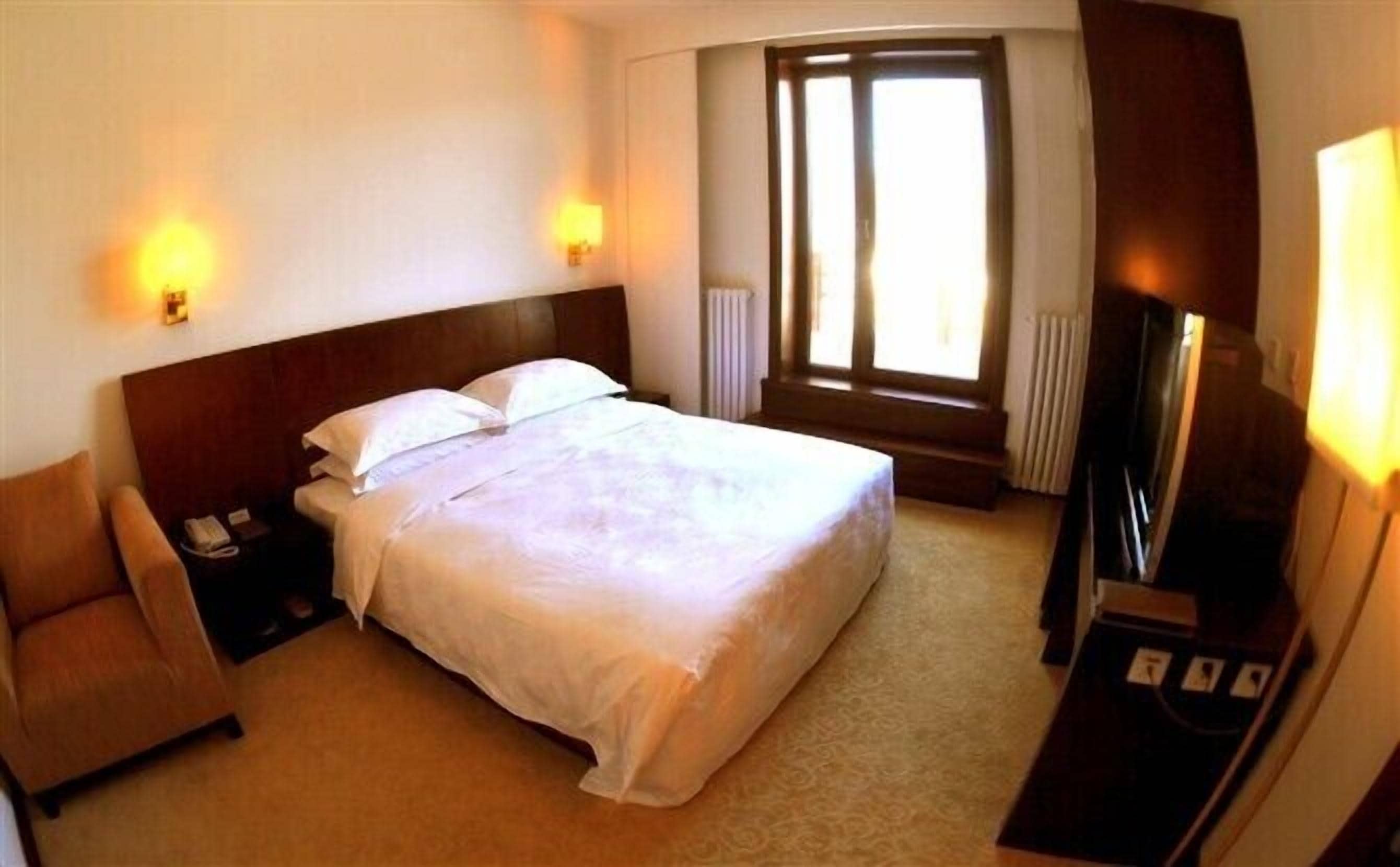 A hotel room with a large bed, bedside lamps, a chair, a desk with a telephone, and a TV.
