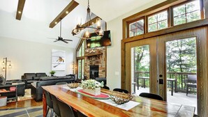 A spacious living area with a large wooden dining table, comfortable seating, a stone fireplace, and large windows offering a view of the outdoors. - Chalet de luxe acceptant les animaux à louer CITQ 240284 (Stoneham-et-Tewkesbury)