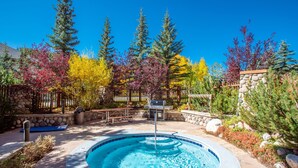 A hot tub surrounded by trees with autumn foliage, a stone wall, and a barbecue grill. - Beautiful Views, Beautiful Condo , wifi, underground park, 1 minute to lifts (Copper Mountain)
