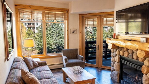 A cozy living room with a sofa, armchair, and coffee table. There is a fireplace with a stone surround and a television above it. Large windows with blinds provide natural light and a view of trees and buildings outside. - Beautiful Views, Beautiful Condo , wifi, underground park, 1 minute to lifts (Copper Mountain)