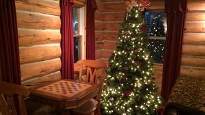 A cozy log cabin interior with a wooden dining table and chairs, a Christmas tree adorned with lights and ornaments, and a window with curtains. - The Gingerbread Cottage - 2.5 acres, craft or meeting room (Bailey)
