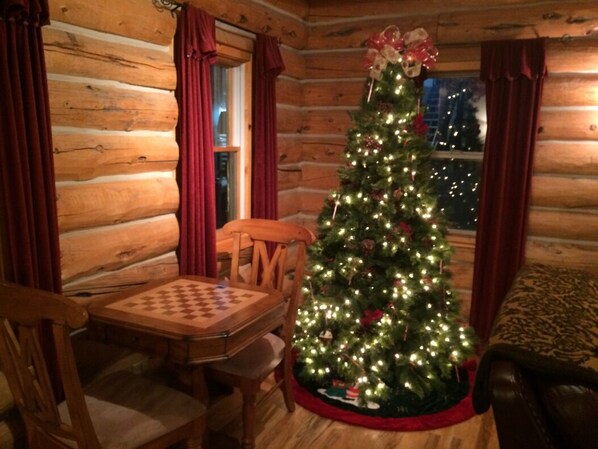L’intérieur d’un chalet confortable, avec des murs en bois, une table et des chaises en bois, un sapin de Noël décoré de guirlandes lumineuses et d’ornements, et une fenêtre avec des rideaux.