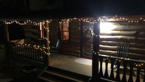 A log cabin with a porch decorated with string lights, a wooden deck, and a small table with chairs. - The Gingerbread Cottage - 2.5 acres, craft or meeting room (Bailey)