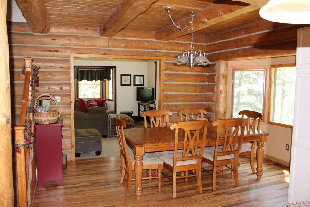 A log cabin interior with a dining table, chairs, a chandelier, a fireplace, and a television in the background.