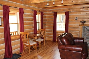 L’intérieur d’une cabane en rondins, avec une table à manger en bois, deux chaises, un canapé en cuir et une cheminée en pierre.