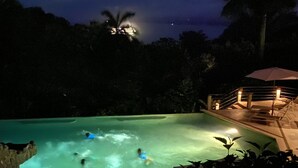 Una piscina nocturna con gente nadando, rodeada de árboles y un área cubierta con luces.