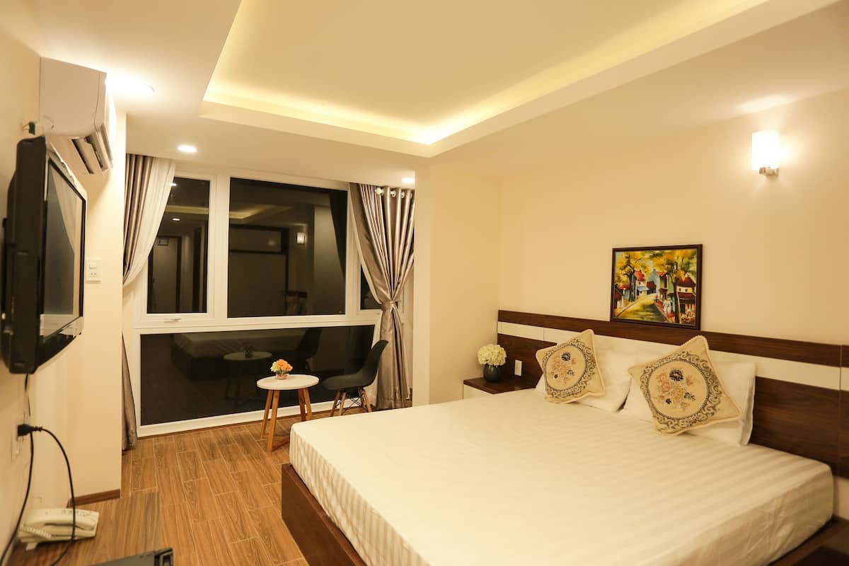 A hotel room with a large bed, a TV mounted on the wall, a small table with a chair, and a window with curtains.