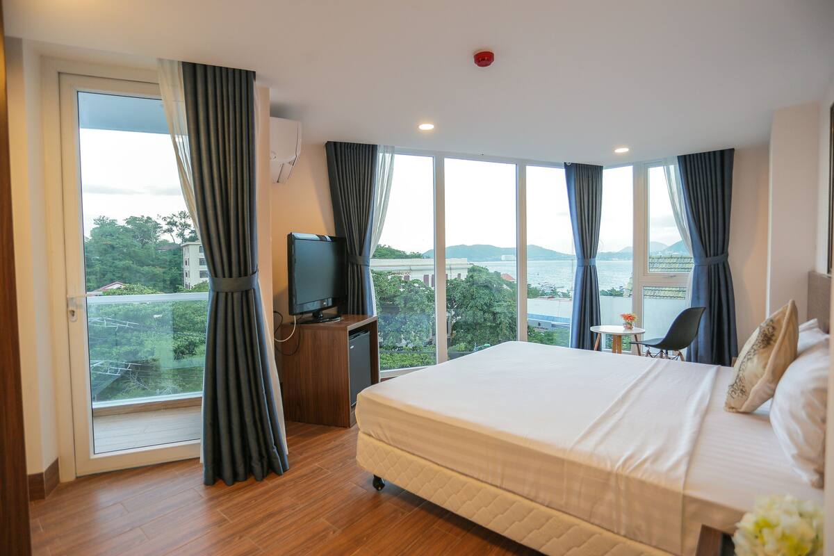 A hotel room with a large bed, a TV, a small table with chairs, and a balcony with a view of the city and hills.