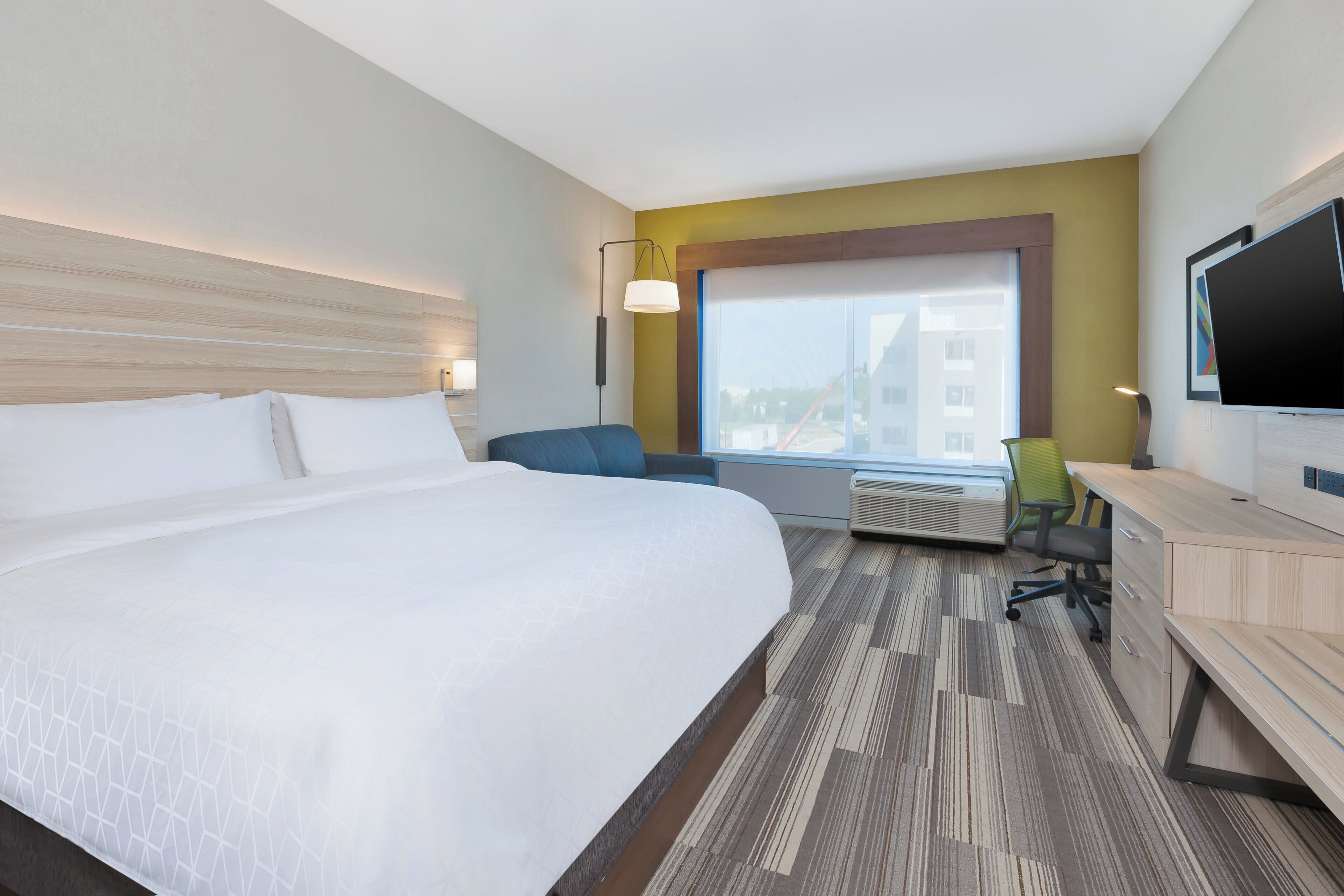 Une chambre d’hôtel avec un grand lit, un bureau avec une chaise, une télévision et une fenêtre donnant sur des immeubles.