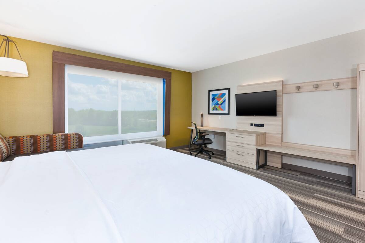 Habitación de hotel con una cama grande, un escritorio con computadora, un televisor y una ventana con vista al exterior.