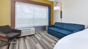 Una habitación de hotel con un ventanal grande, un sofá azul, un sillón gris y una cama blanca.