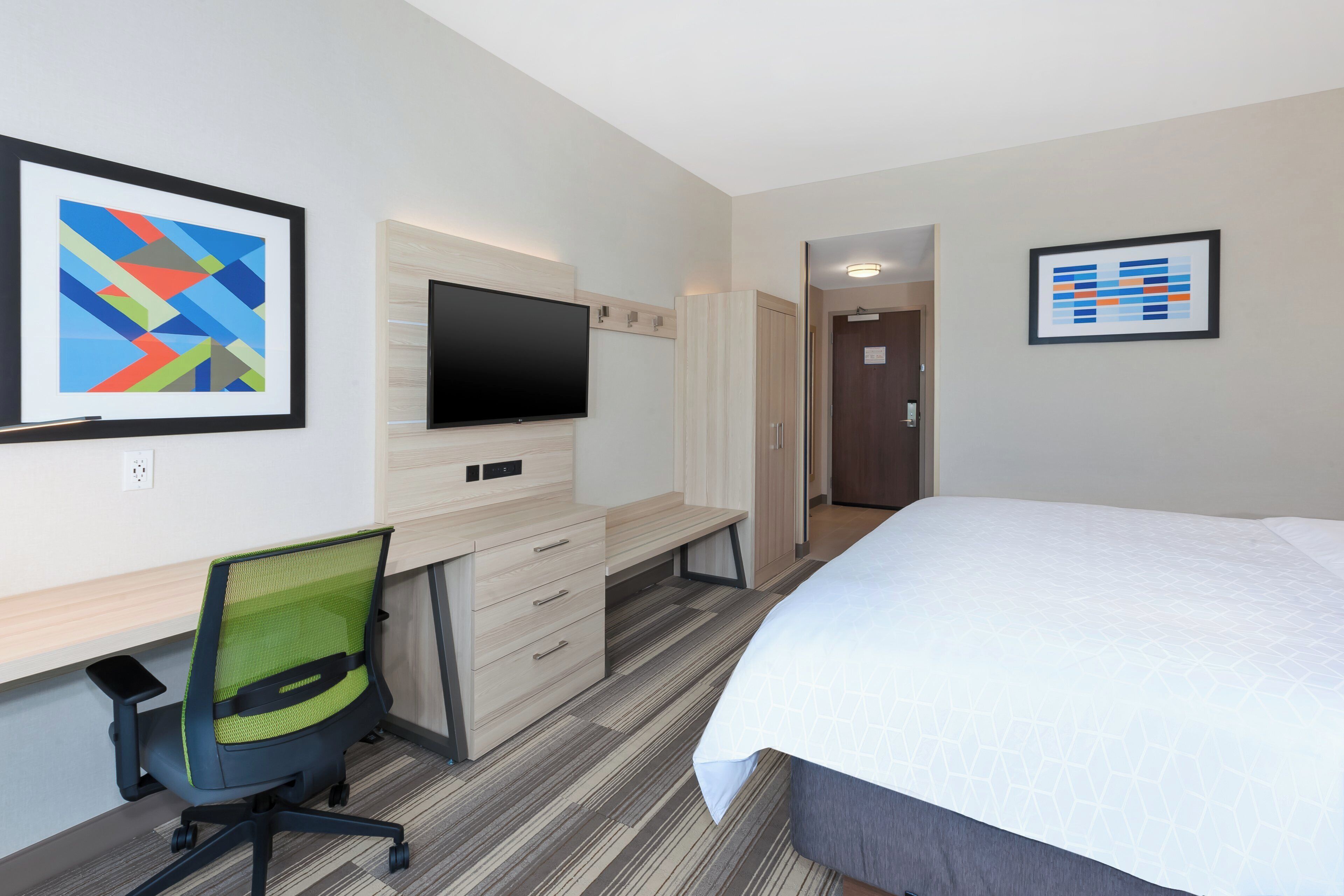 A hotel room with a bed, desk, chair, TV, and artwork on the walls.