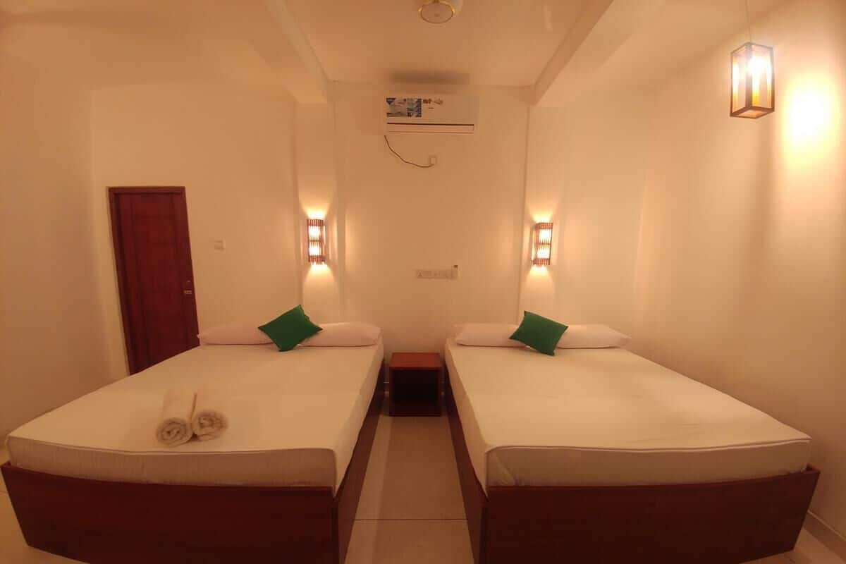 A hotel room with two single beds, a small table, a door, and wall-mounted lights.
