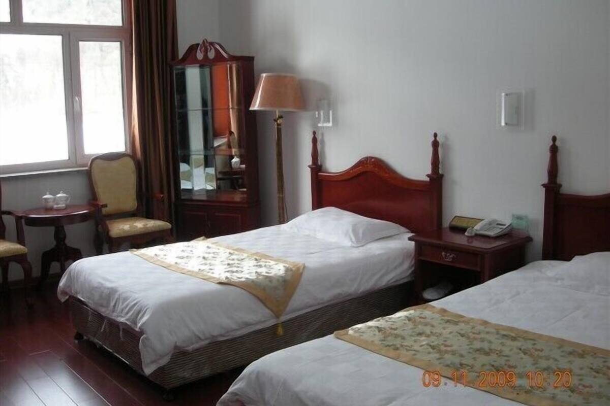A hotel room with two beds, a bedside table with a lamp, a chair, a small table with cups, and a mirror.
