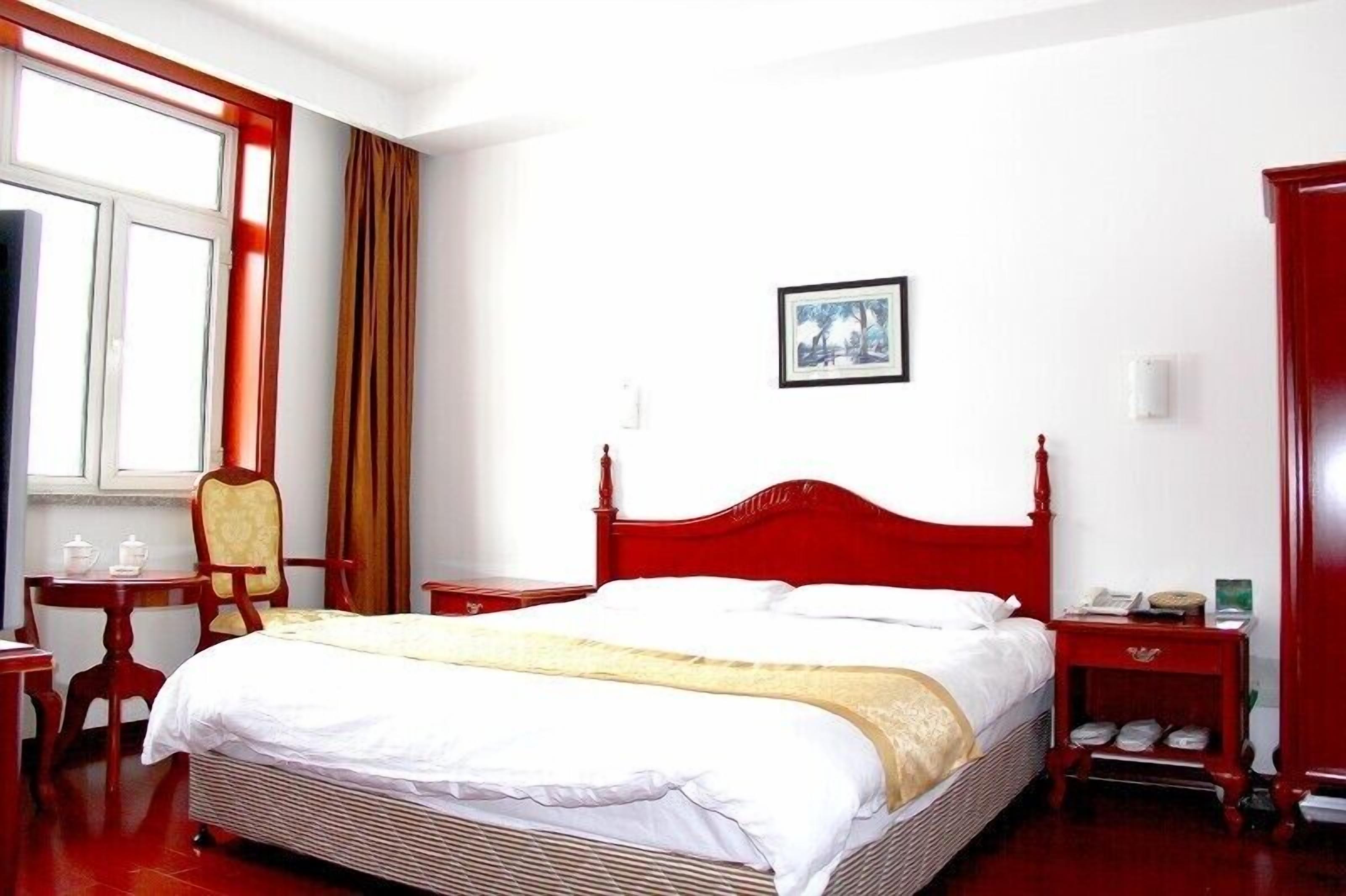 A hotel room with a red bed, a wooden nightstand, a chair, a small table, and a window with curtains.
