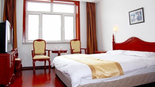 A hotel room with a large bed, two chairs, a small table, a television, and a window with curtains. - Yabuli Olympic Club Hotel (Harbin)