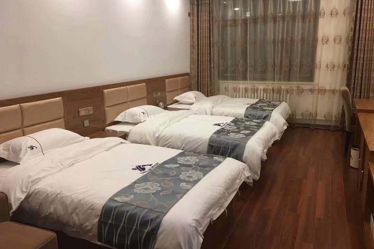 Une chambre d’hôtel avec trois lits, des têtes de lit en bois, un plancher en bois et une fenêtre ornée de rideaux légers.