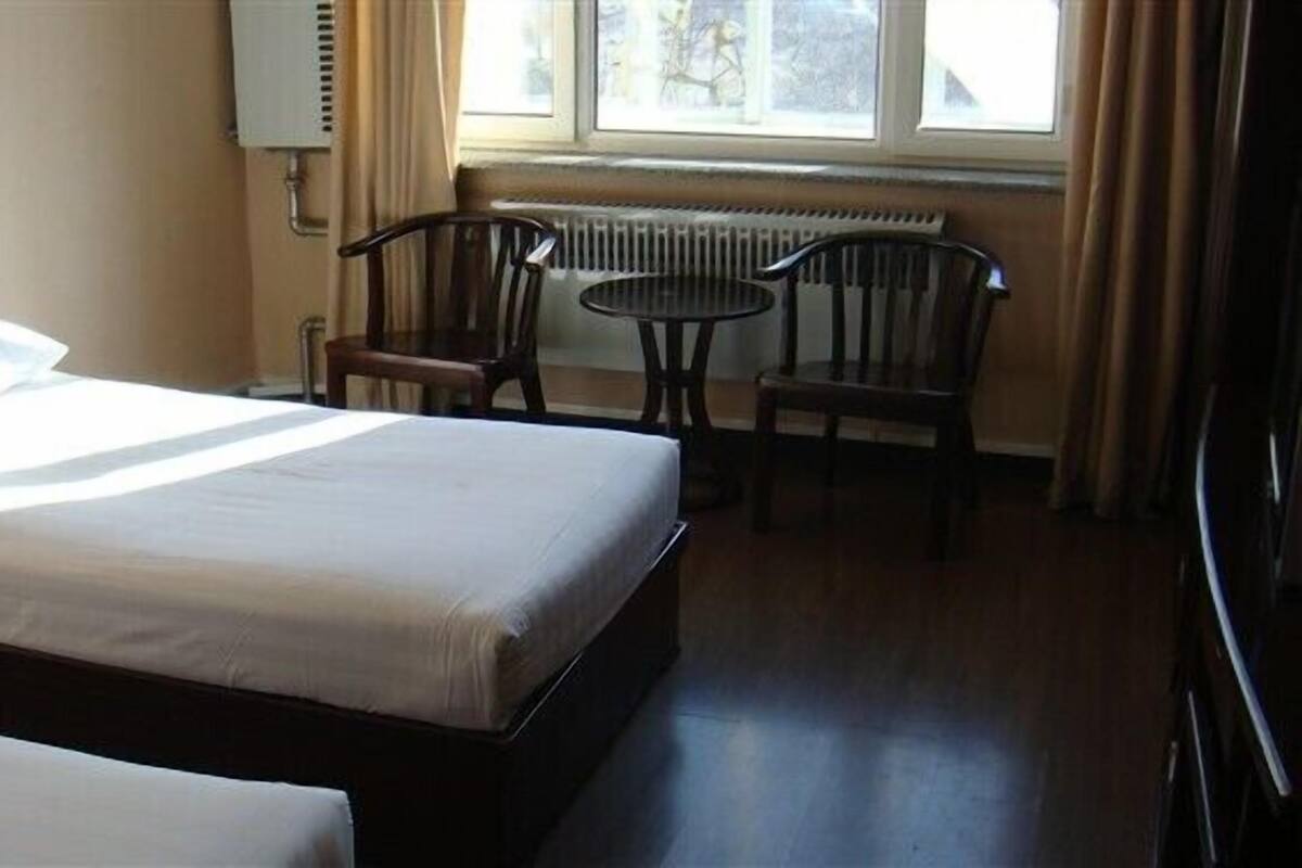 A hotel room with two beds, a small table, two chairs, a radiator, and a window with curtains.