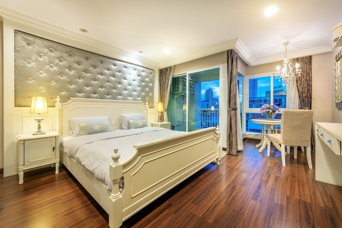 A spacious bedroom with a large bed, a bedside table, a chair, and a view of the city through a large window.