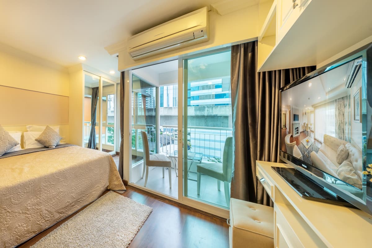 A hotel room with a bed, a TV, a balcony with a view of buildings, and an air conditioning unit.