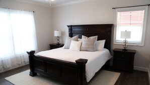 Ein Schlafzimmer mit einem großen Bett, zwei Nachttischen, einem Beistelltisch, einem Fenster mit Jalousien und einer Chandelüstere.