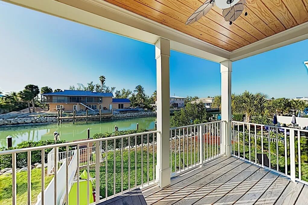 A covered outdoor area with a wooden deck, white railings, and a view of a body of water and buildings in the background.
