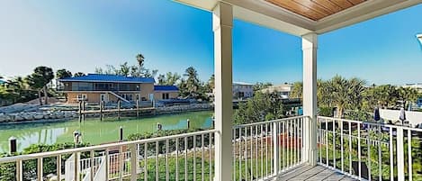 A covered outdoor area with a wooden deck, white railings, and a view of a body of water and buildings in the background.
