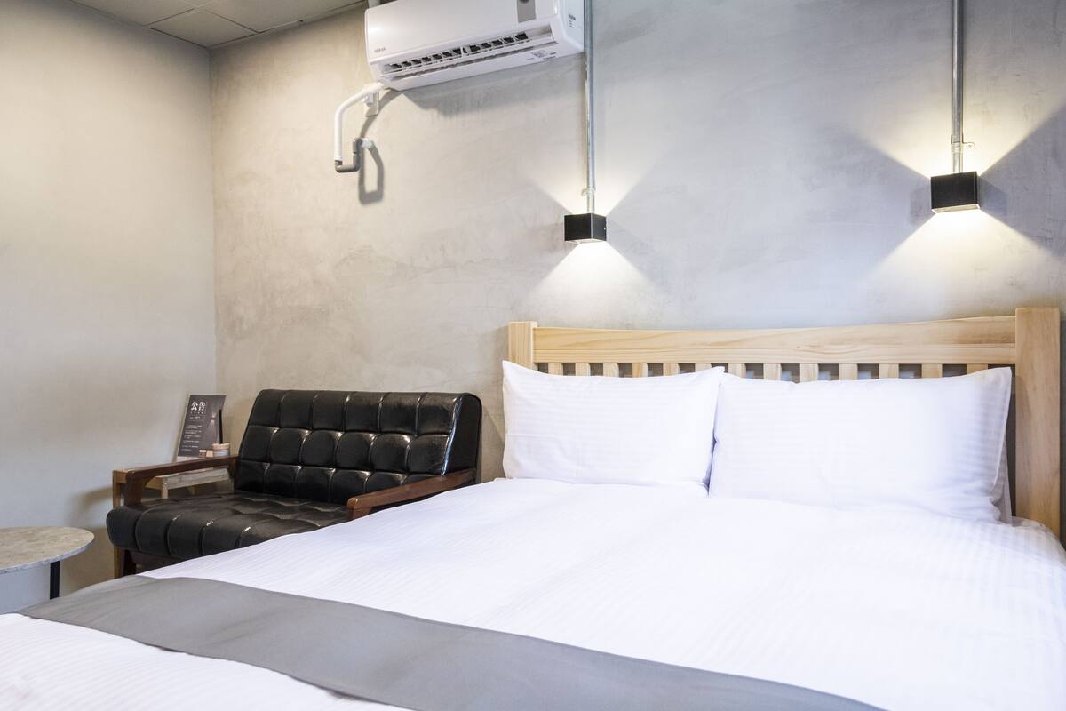 Hotellihuone, jossa on sänky, puinen pääty, musta nahkainen tuoli, pieni pöytä ja seinälle asennettu ilmastointilaite.