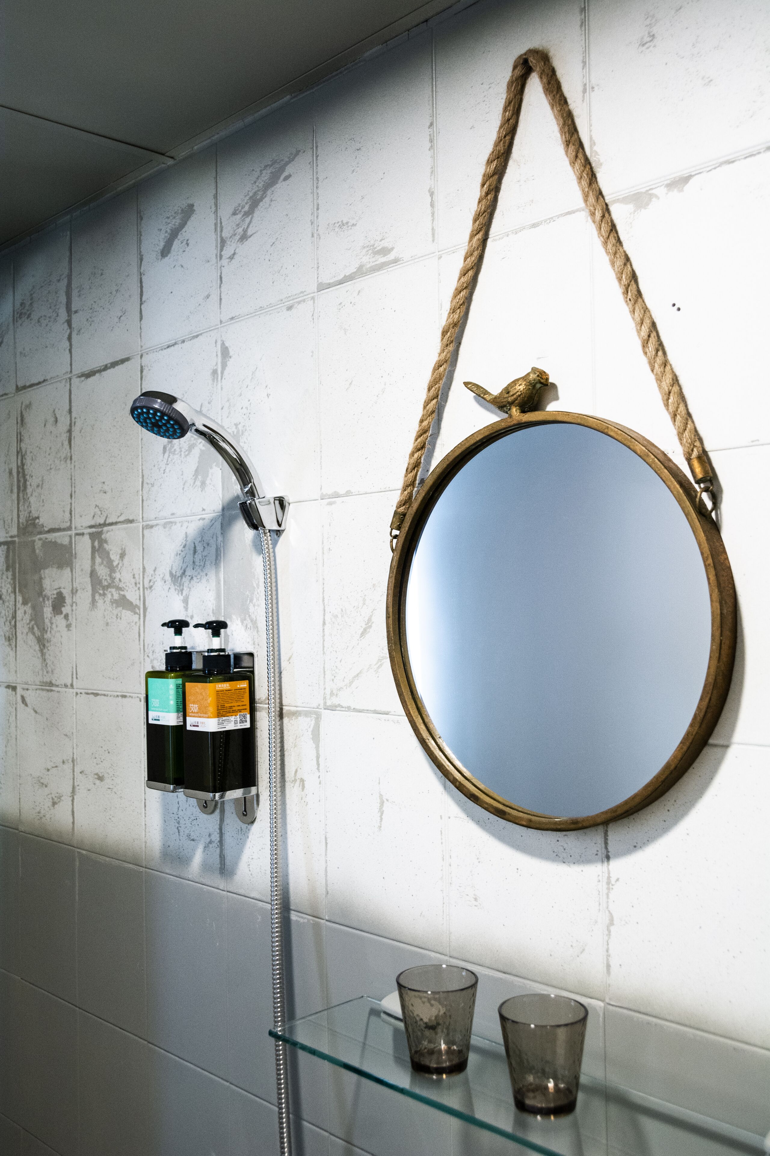 Un baño con ducha, un espejo redondo con asa de cuerda y dos vasos de vidrio en una estantería.