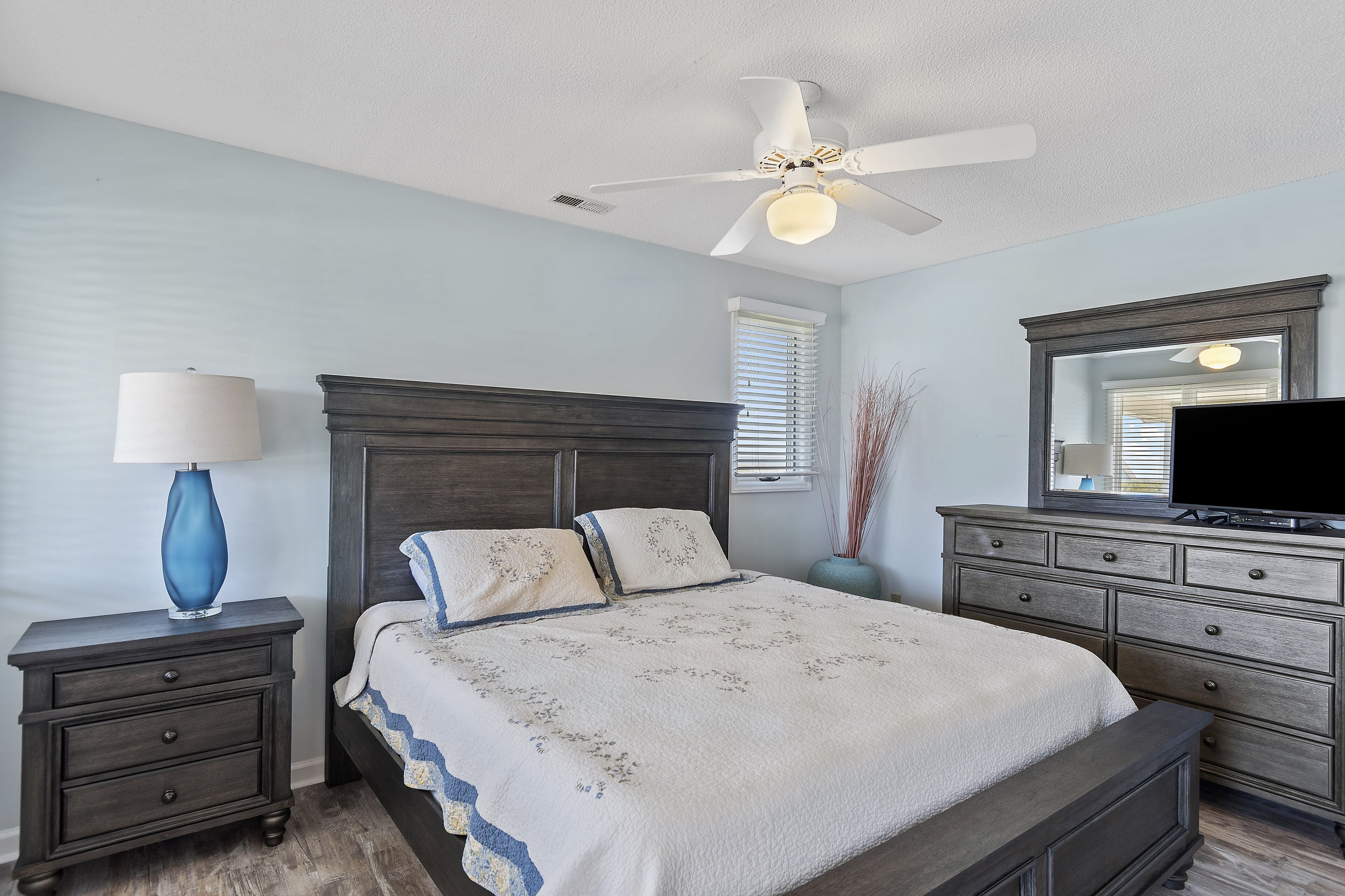 A bedroom with a bed, nightstand, dresser, ceiling fan, and a television.