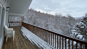 Una terraza de madera con baranda, sillas de plástico blanco y vista a árboles y colinas cubiertas de nieve.