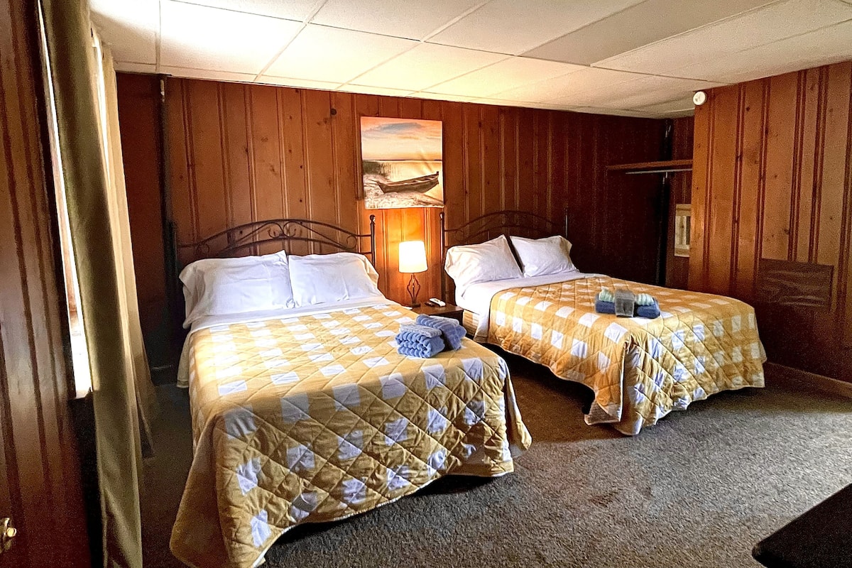 A room with two beds, wooden walls, a painting, and a nightstand with a lamp.