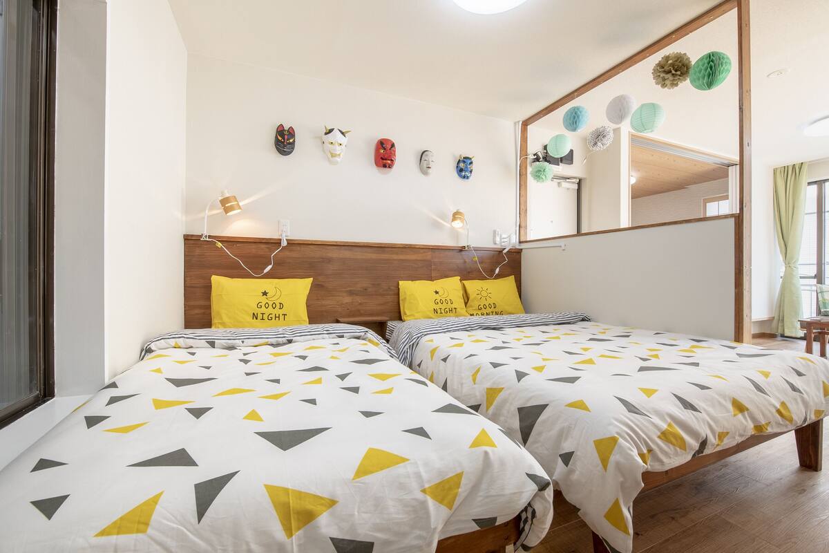 Une chambre avec deux lits, chacun agrémenté d’une couvre-lit à motifs géométriques, d’une tête de lit en bois et de deux oreillers jaunes ornés de motifs textuels.