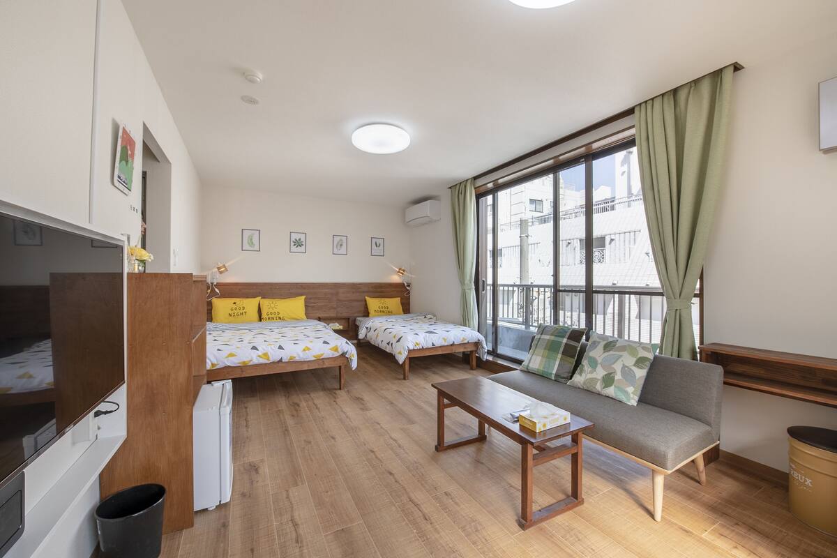 Une chambre avec deux lits simples, un canapé, une petite table, une télévision et une fenêtre avec des rideaux.