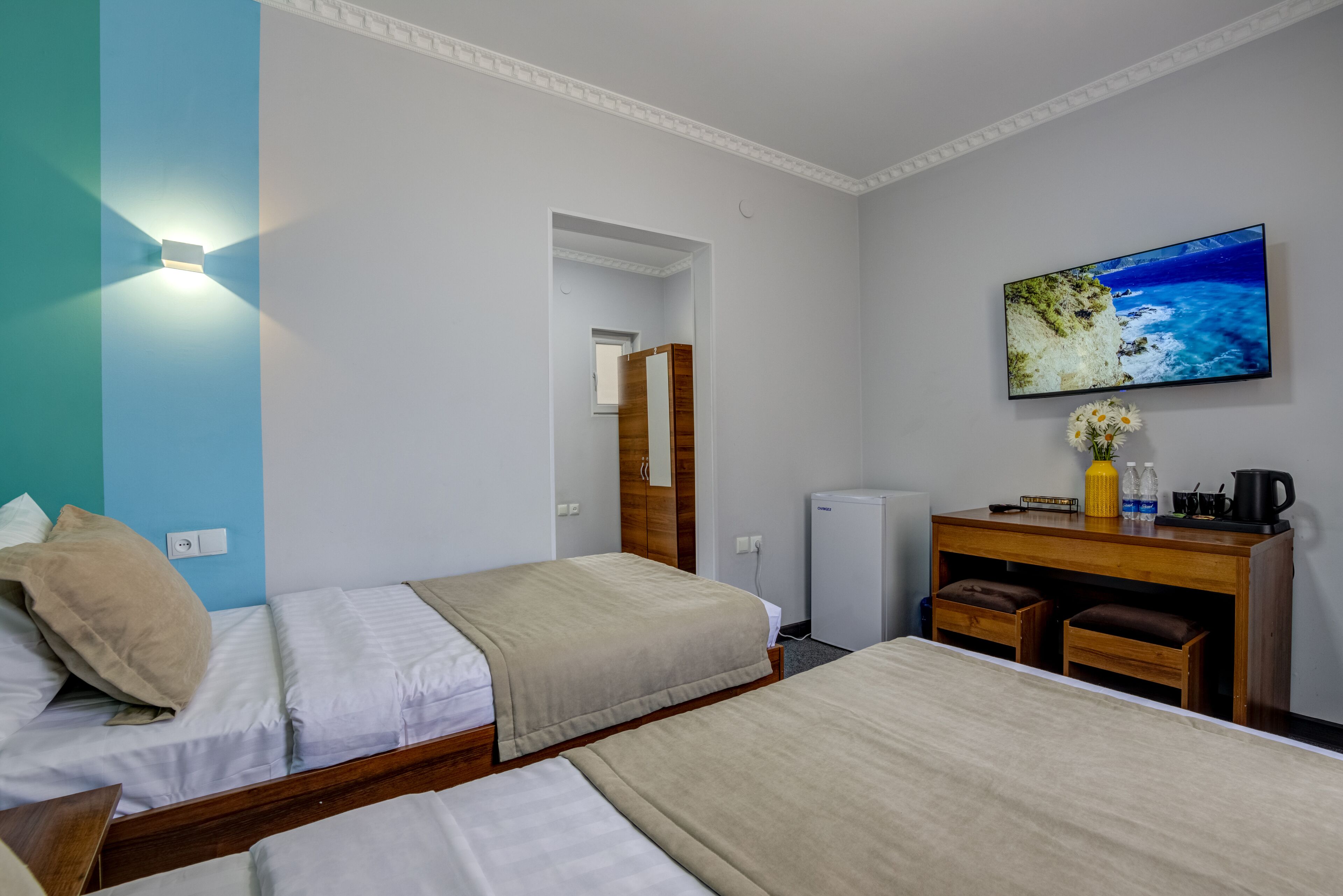 A hotel room with two single beds, a wooden dresser with a mirror, a small refrigerator, and a television mounted on the wall displaying a scenic ocean view.