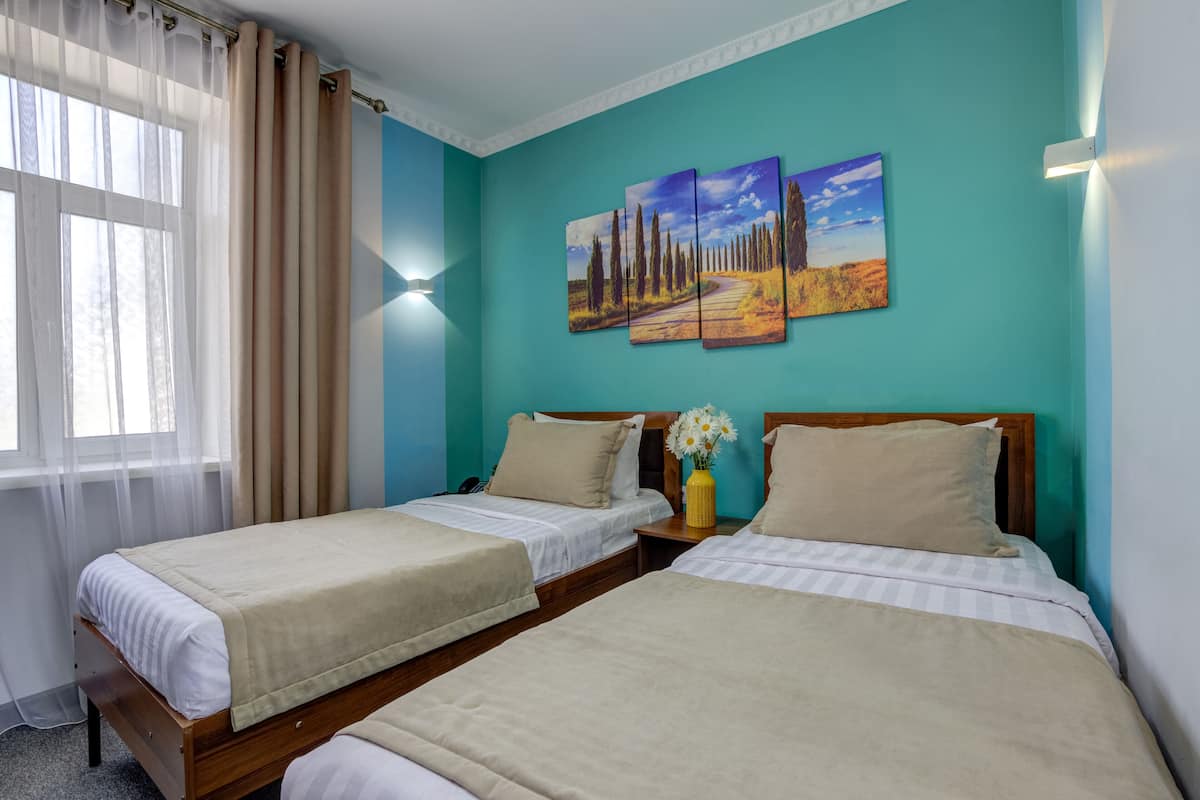 Una habitación con dos camas, una ventana con cortinas, una mesita de noche con un florero y una pared con tres cuadros.